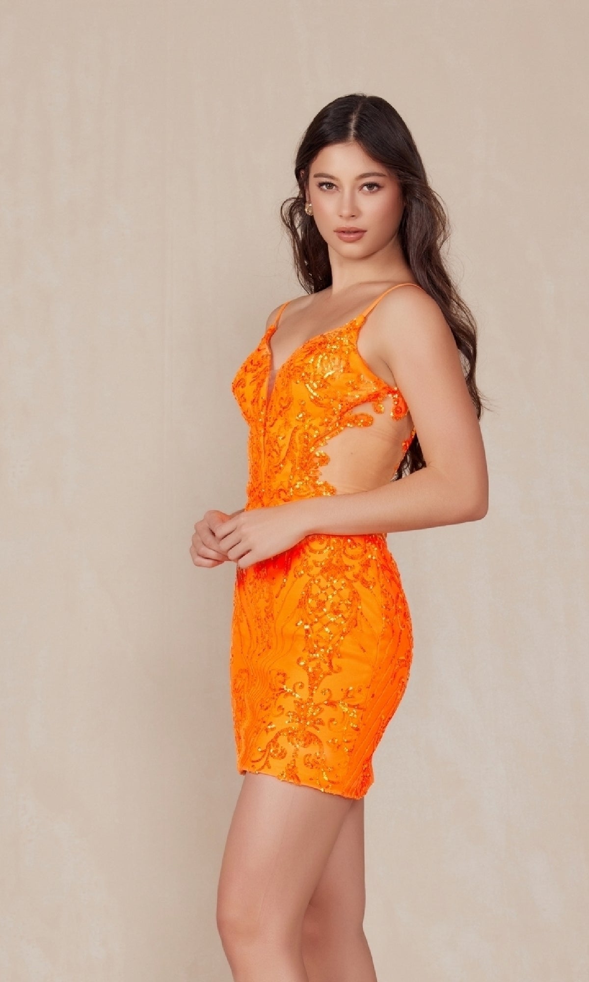 A woman with long dark hair models the Sheer-Sides Short Sequin-Print Hoco Dress D852, a sleeveless, bright orange cocktail dress with side cutouts and sequins, standing against a plain beige background.